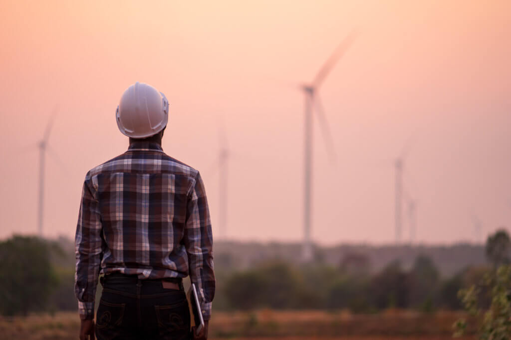 An engineer from behind, carrying a tablet and staring at wind turbines in the distance.