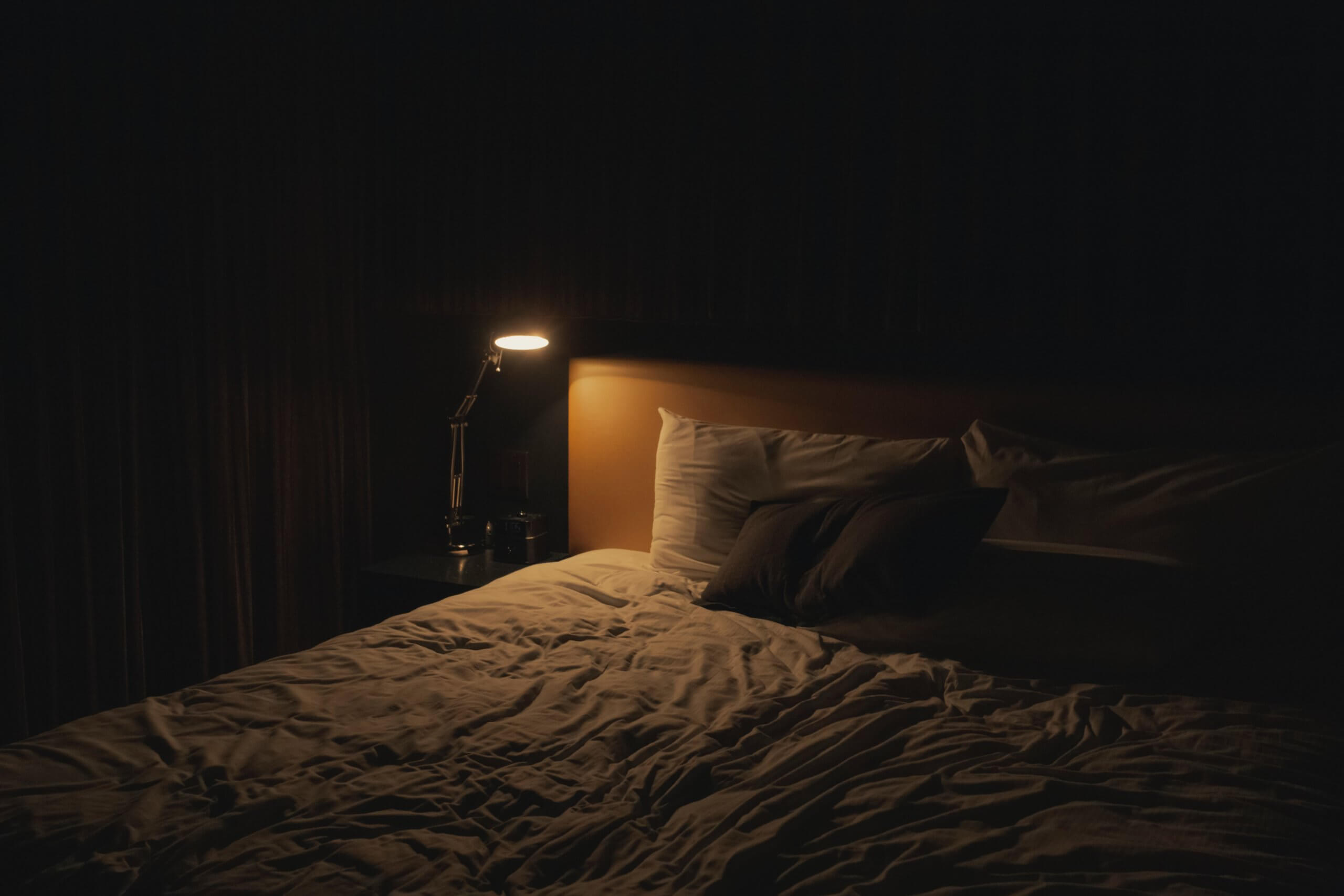 A lamp on a bedside table illuminating the corner of a bed.